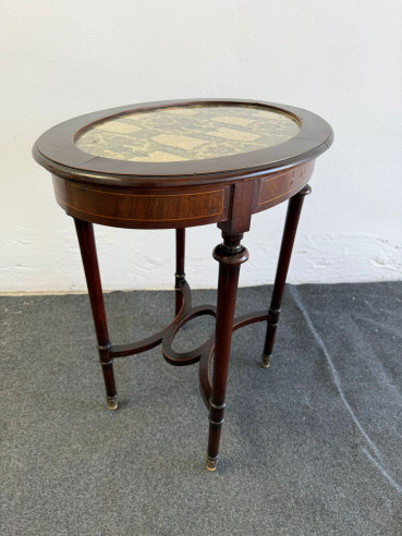 Elegante mesa auxiliar, ovalada, en caoba con cristal y tapizado bordado de fondo.